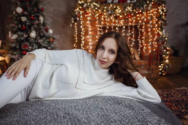 居心地の良いセーターで完璧なボディを持つ美しいモデルの女の子は 新年のために装飾されたインテリアのベッドの上に座っています 若いです女性でModish居心地の良い服Posingオンザベッドに対してクリスマスツリーでザロフト — ストック写真
