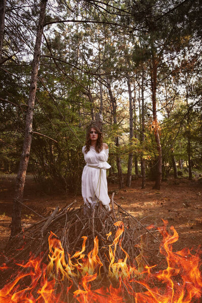 Beautiful mystery gothic woman in long white dress in autumn forest. Dark fantasy creature, bonfire for a witch