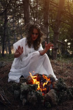 Sonbahar ormanında uzun beyaz elbiseli, gizemli gotik kadın. Karanlık fantezi yaratığı. Cadı ateşin yanında oturuyor.