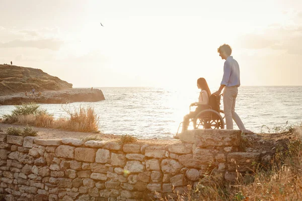 redhair woman in wheelchair talking with her partner by the sea at sunset. Summer tropical beach vacation.