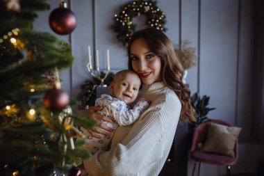 Beyaz kazaklı anne küçük çocuğu öpüyor Noel ağacındaki süveterli çocuğu ve evdeki çelenkleri.