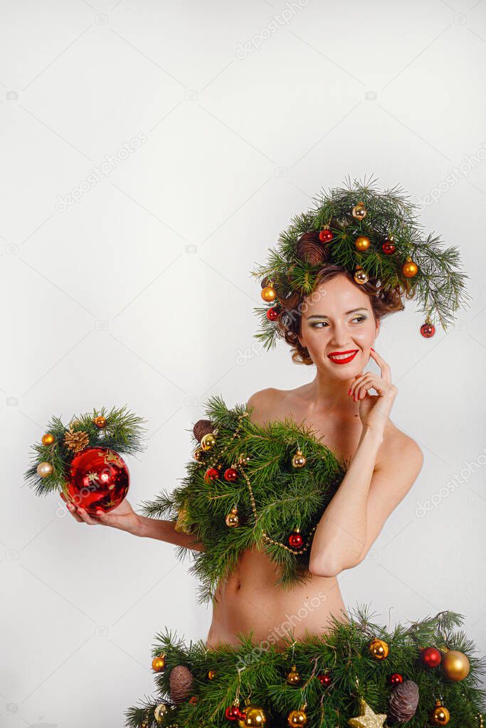 Beautiful happy young lady with a Christmas pine wreath on her head and fashione xmas tree dress, decorated with Christmas balls, berries and snowflakes. New Year Concept
