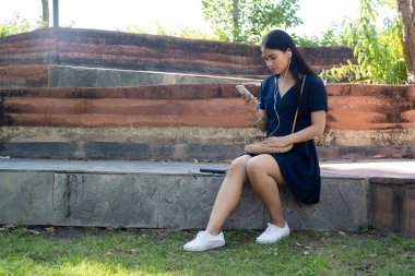 Koyu mavi elbiseli uzun saçlı kadın erkek arkadaşını beklerken parktaki bankta çalan müziği dinle..