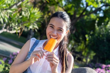 Kot pantolonlu, beyaz tişörtlü, atkuyruklu kız güneşli bir günde parkta portakallı dondurmayı ısır. Yüzünde mutluluk gülümsemesi var..