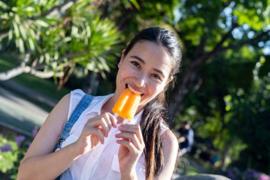 Kot pantolonlu, beyaz tişörtlü at kuyruklu kız iki eliyle dondurmayı kavrayıp kameraya gülümserken ısır..
