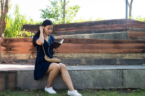 公園のベンチに座っている濃い青のドレスの長い髪の女性は 彼女のボーイフレンドを待っている本を読んで楽しむ 彼女はまた イヤホンで曲を聞く — ストック写真