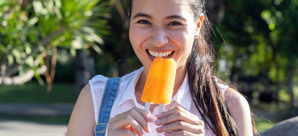 Ponytail girl in denim overalls and white shirt bite an orange ice cream in the park in sunny day, she has the smile of happiness on her face.