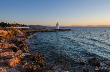 Aegina adasının vahşi kıyıları ve arka plandaki eski deniz feneri, Saronik Körfezi, Yunanistan, gün batımında.