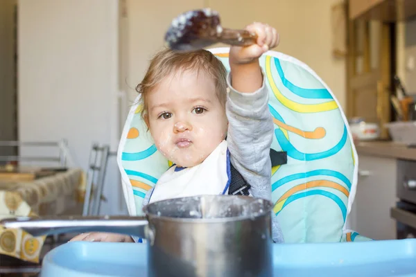 Funny baby child getting messy eating cereals or porridge by itself with a wooden spoon, straight from the cooking pot