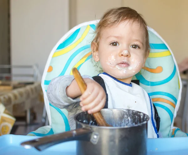 Funny baby child getting messy eating cereals or porridge by itself with a wooden spoon, straight from the cooking pot