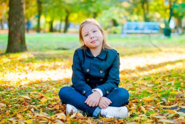 Komik küçük yürümeye başlayan çocuk kız mavi bir ceket ve kot bir güneşli parkta altın yaprakları sarı ve portakal ağaçları ile sıcak bir sonbahar günü oturan 