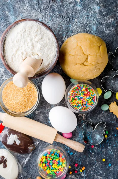 Easter cooking background, dough and indredients, cuts for cookies and colored sugar decor for baking on dark background, The process of making fragrant Easter cookies, series photo, top view, flat lay, copy space