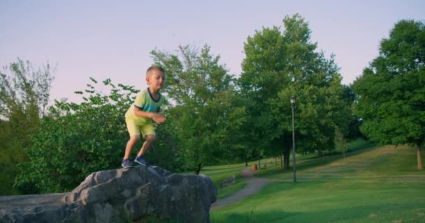 Netter kleiner Junge, der im Sommerpark vom Felsen springt. Rote Kinokamera. Zeitlupe 4k — Stockvideo