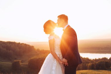 Gelin ve damat el ele tutuşup doğa günbatımı sırasında öpmek