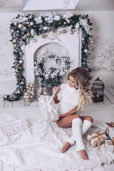 一个穿着白色衣服坐在地板上的女孩打开圣诞礼物 昂贵的内饰 白色装饰为新年假期 — 图库照片