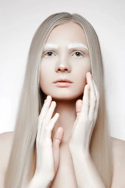 Portrait of a young girl with blond hair. White makeup on the eyebrows. Girl touches face with her hands