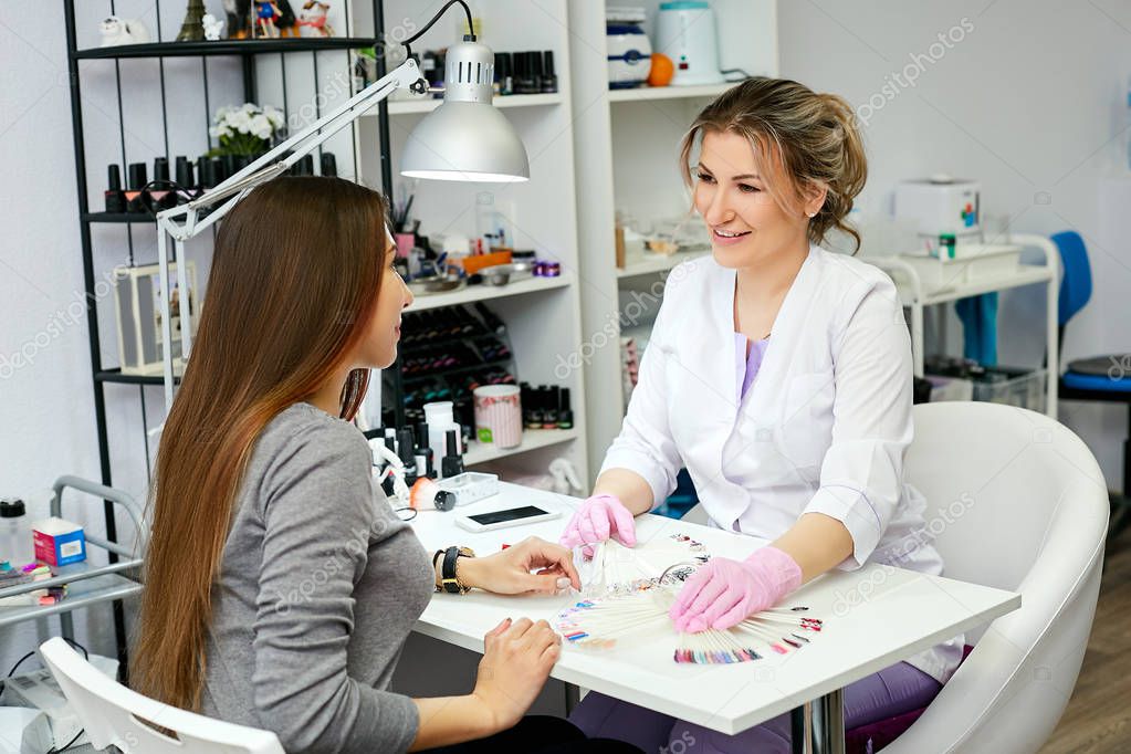 A brunette girl in a gray sweater consults a manicurist and a nail artist
