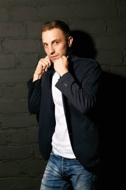 Yakışıklı delikanlı mavi ceket ve kot pantolon ve gri tuğla duvara standları yakasını düzeltiyor