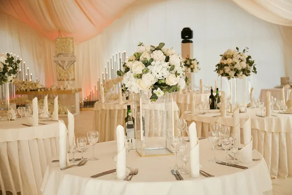 Dining tables decorated with bouquets of white flowers in the tent during the wedding ceremony.