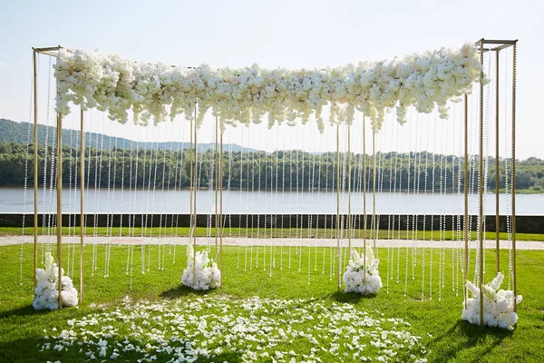 豪華なアーチは 屋外の川の背景の下で新婚の出口登録時に白い花の花束で飾られました 芝生の白いバラの花びらで覆われています ストック画像