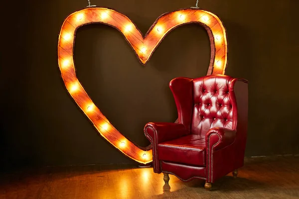 Uma luxuosa poltrona vermelha feita de couro genuíno contra o fundo uma parede marrom. Um coração de madeira com luzes no fundo — Fotografia de Stock