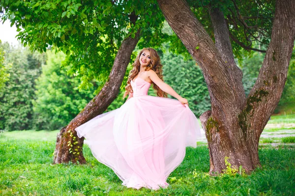 Молодая девушка в розовом платье кружится и улыбается в летнем парке — стоковое фото