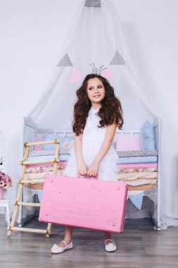 Başında bir taç ile mutlu bir kız, beyaz bir elbise ve ayakkabı yumuşak renklerde çok renkli şilte bir sürü bir çocuk yatağının arka planına karşı pembe bir bavul tutuyor.