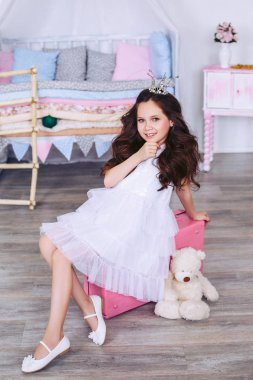 Beyaz elbiseli gür saçlı bir kız, elini çenesinde tutarak, oyuncak bej ayı ile pembe bir bavul üzerinde oturuyor.