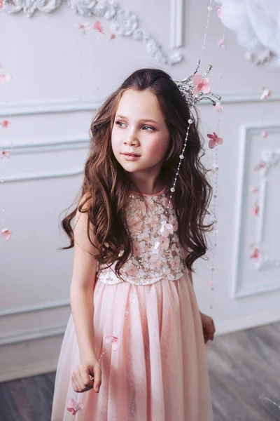 白い古典的な壁に対する夢らしい表情を持つピンクのドレスを着た小さな黒髪の女の子は、彼女の頭の上に王冠を持つピンクのドレスに立っています. — ストック写真