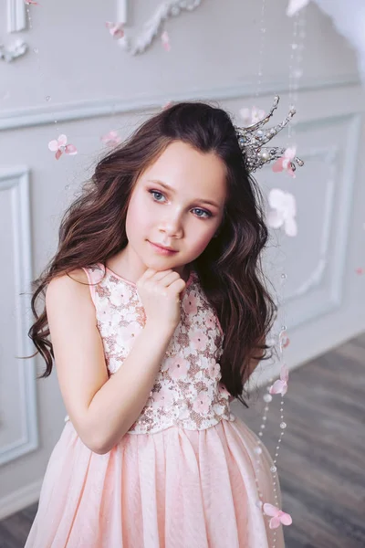 美しいピンクのドレスと彼女の頭の上に王冠で白い壁に対してポーズ夢の表情を持つかわいい女の子. — ストック写真
