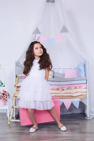 一个穿着浅色连衣裙的女孩手里拿着一个粉红色的手提箱，面带微笑地看着。在儿童床的背景. — 图库照片