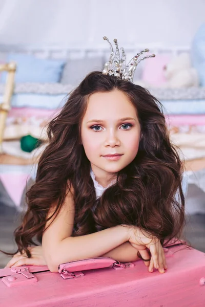 Retrato de una niña con rizos lujosos con una corona de piedras y perlas en la cabeza apoyada en una maleta rosa. La chica en la imagen de una princesa en un guisante . — Foto de Stock