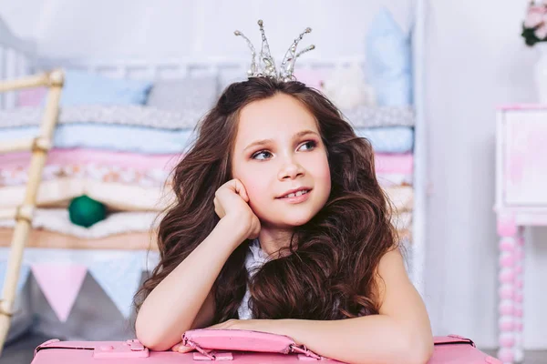 The little girl with thick dark hair and a crown on her head dreamily looks away leaning on a pink suitcase. — Stock Photo, Image