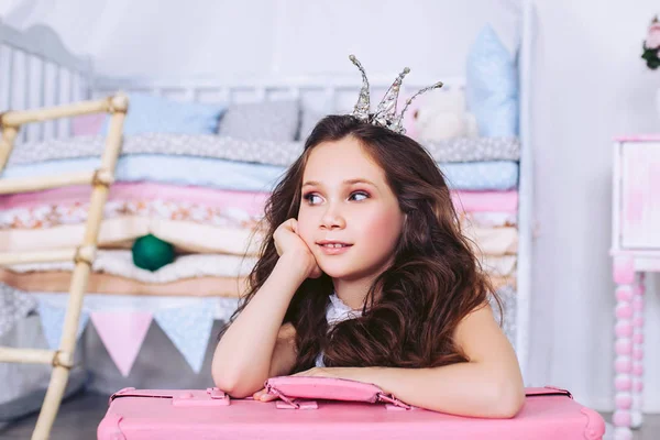 Een schattig meisje met donker haar en een kroon op haar hoofd ziet er weg uit te grijpen in een mooie roze koffer. — Stockfoto