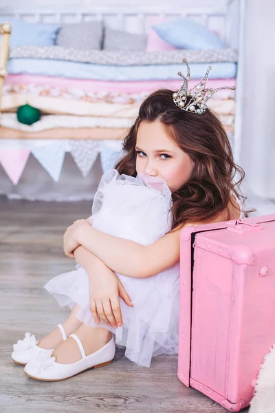 Uma menina bonita em um vestido branco bonito e uma coroa em sua cabeça está sentada no chão com as pernas cruzadas com as mãos ao lado de uma mala rosa . — Fotografia de Stock
