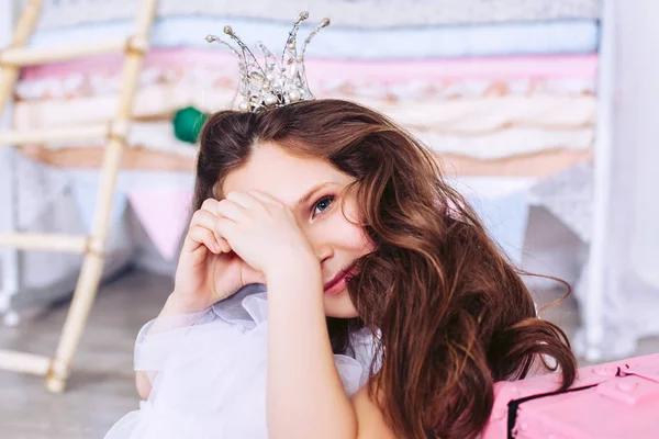 Маленькая девочка с короной на голове закрывает один глаз рукой, сидя в детской комнате рядом с розовым чемоданом. . — стоковое фото
