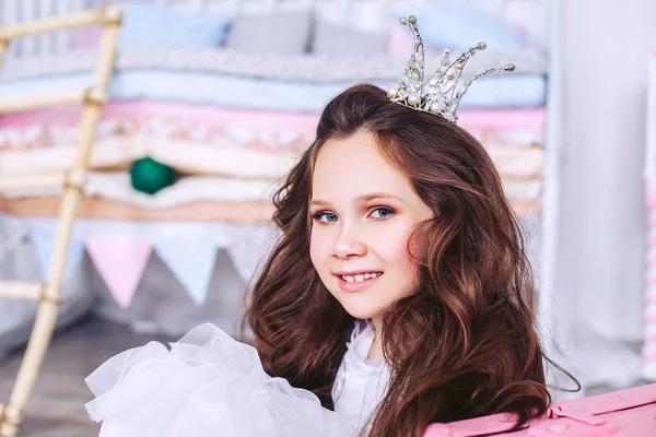 Een klein meisje met een kroon op haar hoofd en in een witte jurk zittend op de vloer in de kinderkamer ziet er recht aan haar hoofd aan de zijkant. — Stockfoto