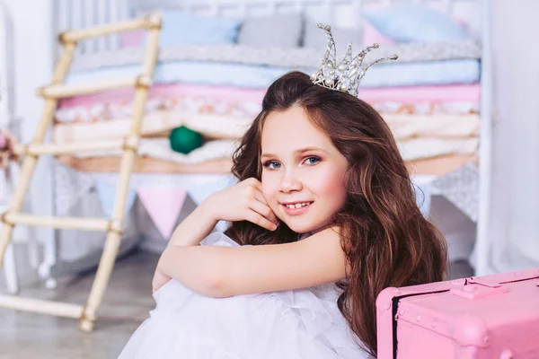 Una niña pequeña con una corona en la cabeza posando en el vivero junto a una maleta rosa . — Foto de Stock