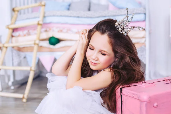 Uma menina em um lindo vestido branco com uma coroa na cabeça está sentada no chão com os olhos fechados e sorrindo. Menina sonhadora . — Fotografia de Stock