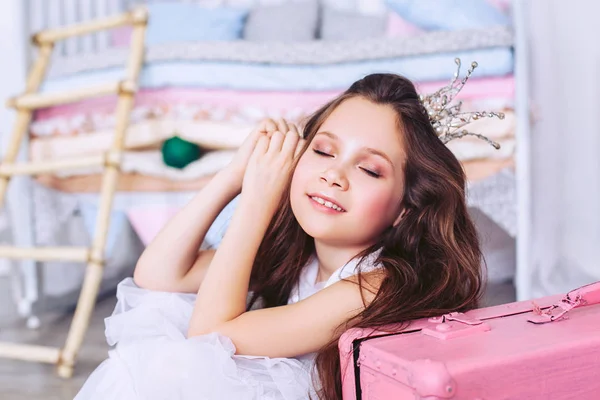 En söt liten flicka med slutna ögon sitter på golvet bredvid en rosa resväska och leende. En flicka med en krona på huvudet. — Stockfoto