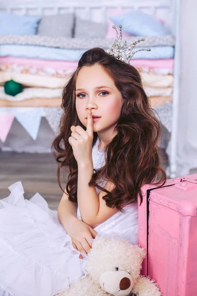 Una bambina con un bel vestito bianco e una corona sulla testa mostra un segno di silenzio seduta sul pavimento accanto a una valigia rosa e un peluche . — Foto Stock