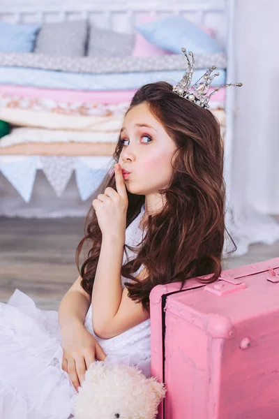 Uma menina com uma coroa na cabeça está brincando enquanto sentada no chão em um vestido branco ao lado de uma mala rosa . — Fotografia de Stock