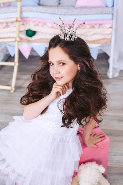 Hermosa niña con el pelo largo y oscuro posa con una corona en la cabeza en el vivero. Lindo bebé en un vestido blanco de lujo . — Foto de Stock