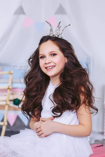 Una hermosa niña con lujoso cabello oscuro y una corona en la cabeza sonríe desafiantemente y posa durante una fiesta de cumpleaños. Bebé feliz en un vestido blanco . — Foto de Stock