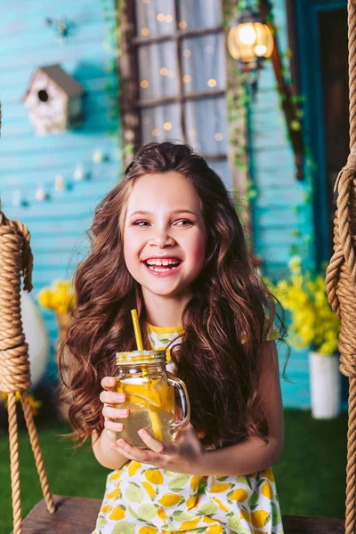 Pequena menina de cabelos escuros ri sentado em um balanço, segurando limonada em suas mãos — Fotografia de Stock
