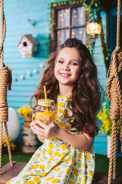 A girl is smiling while sitting on a children's swing with lemonade in her hands. — Stock Photo, Image