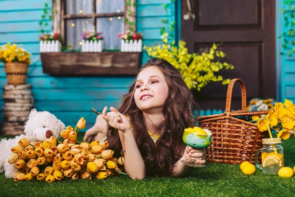 黄色い花の中の豪華な女の子は、家の近くのピクニックで黄色と緑のアイスクリームとスプーンを持って、離れて見て笑顔. — ストック写真