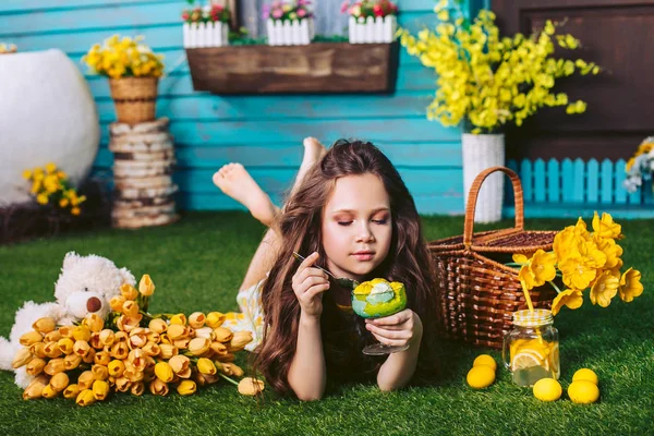 Милая девушка лежит на зеленой лужайке, ест желто-зеленое мороженое с ложкой рядом со светлым плюшевым мишкой, корзинкой для пикника, желтыми цветами, лимонадом и пасхальными яйцами . — стоковое фото