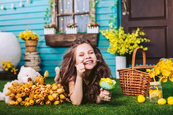 Прекрасная веселая девушка с ложкой мороженого в зубах, лежащая на траве на пикнике на фоне синего дома . — стоковое фото