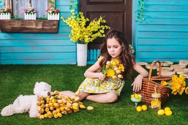 Маленькая девочка сидит на лужайке возле дома, держа в руках букет жёлтых цветов, опираясь на корзину для пикника. Болезненный взгляд ребенка . — стоковое фото